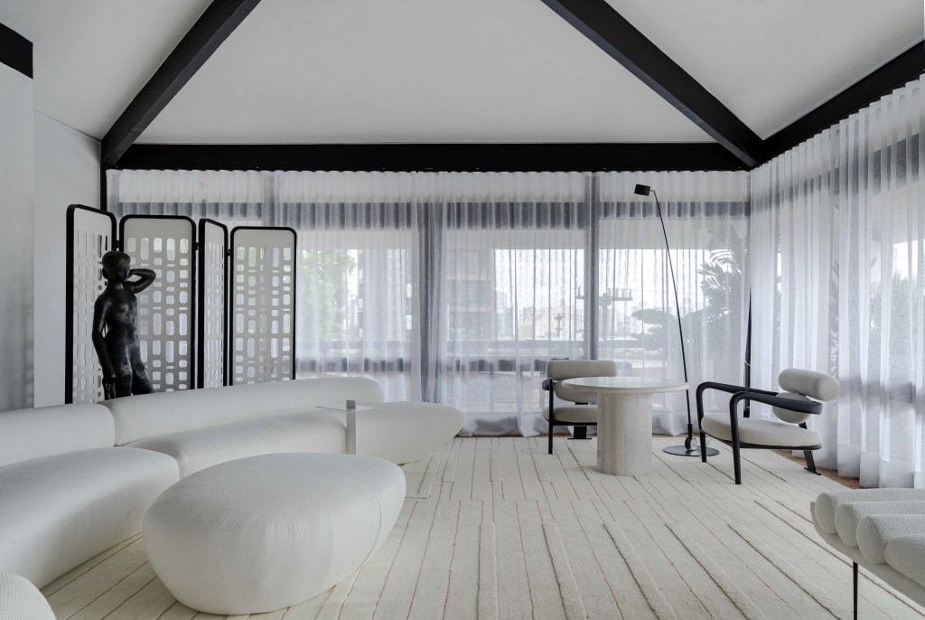 Living minimalista de 85 m² ganha décor em branco, preto, cinza e areia. Projeto de David Bastos para a CASACOR Bahia. Na foto, sala de estar com sofá curvo, estante preta, espelho e tapete.