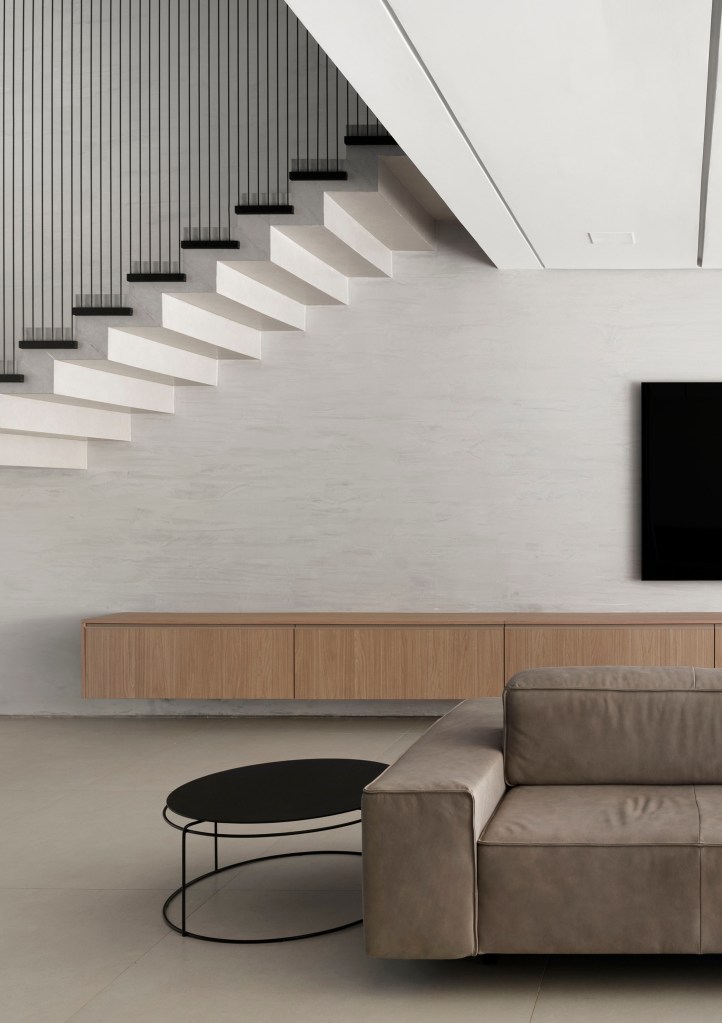 Sala de tv minimalista com parede cinza clara, rack de madeira e sofá baixo; escada branca com guarda corpo preto.
