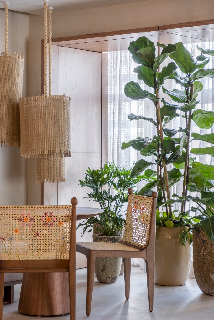 Sala de estar em tons beges, mesa redonda pequena de madeira, cadeiras de palhinha, luminária de palhinha e plantas.