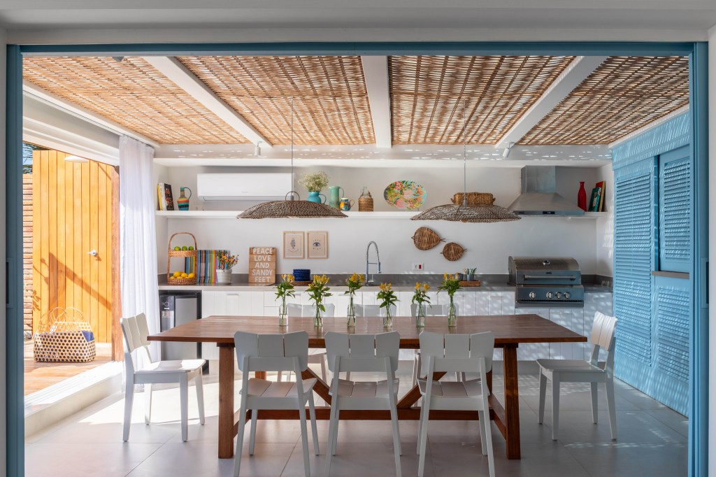 casa-de-praia-inspiracao-grega-area-gourmet-24m-brise-arquitetura-05 Projeto de Brise Arquitetura. Na foto, área gourmet com cobertura de pergolado com palhinha, mesa de madeira, cadeiras brancas e bancada de cozinha.