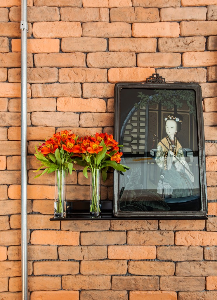 Projeto de Korman Arquitetos. Na foto, parede de tijolinhos rústicos com prateleira suspensa preta com quadro e vasos de flores.