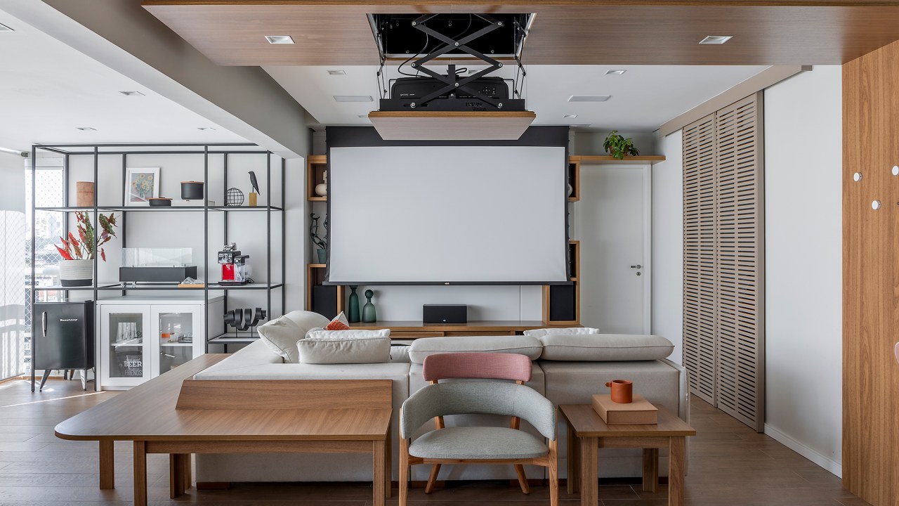 Telão embutido no forro da sala é destaque deste apê de 130 m². Projeto de Ju Matos Arquitetura. Na foto, sala de estar com tv, estante vazada e sofá em L. Telão embutido no forro.