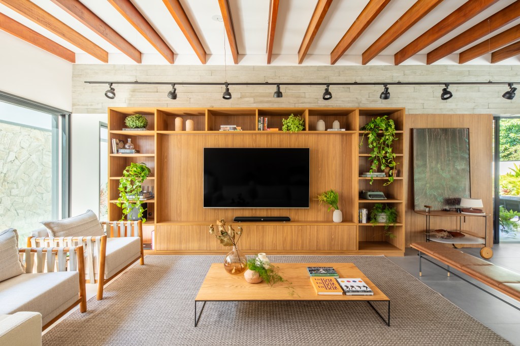 Com sala de jogos e área gourmet ampla, casa de campo vira lar permanente. Projeto de Pietro Terlizzi Arquitetura. Na foto, sala de estar, painel de madeira com estante de nichos, mesa de centro de madeira.