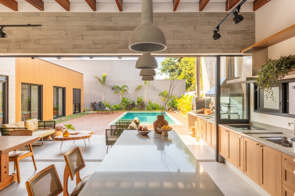 Com sala de jogos e área gourmet ampla, casa de campo vira lar permanente. Projeto de Pietro Terlizzi Arquitetura. Na foto, área gourmet com bancada branca, banquetas de madeira e piscina ao fundo.