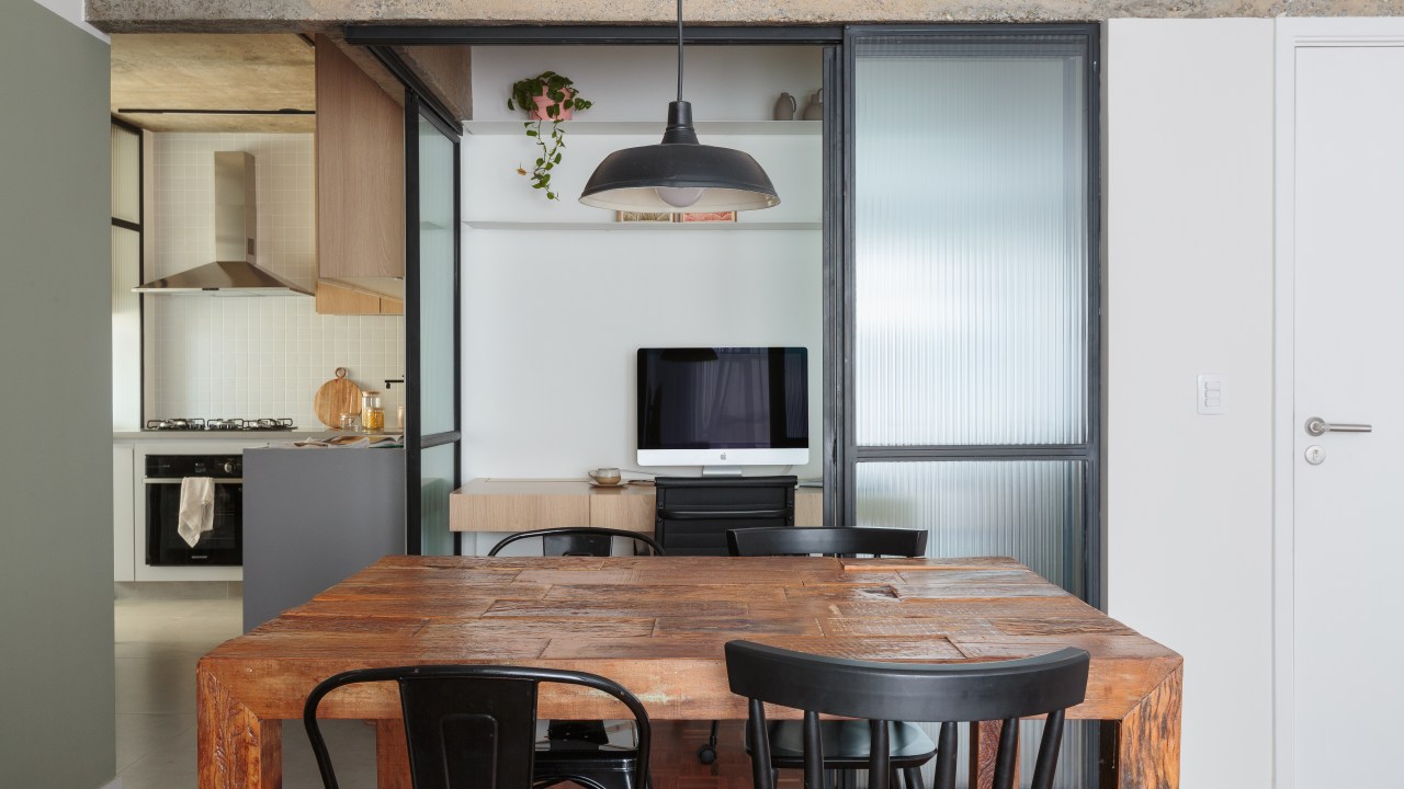 Apê de 70 m² ganha toque industrial com viga aparente e cama em metal. Projeto de Estúdio Maré. Na foto, sala de jantar com mesa de madeira, cadeiras pretas, home office com porta de vidro canelada, cozinha ao fundo.