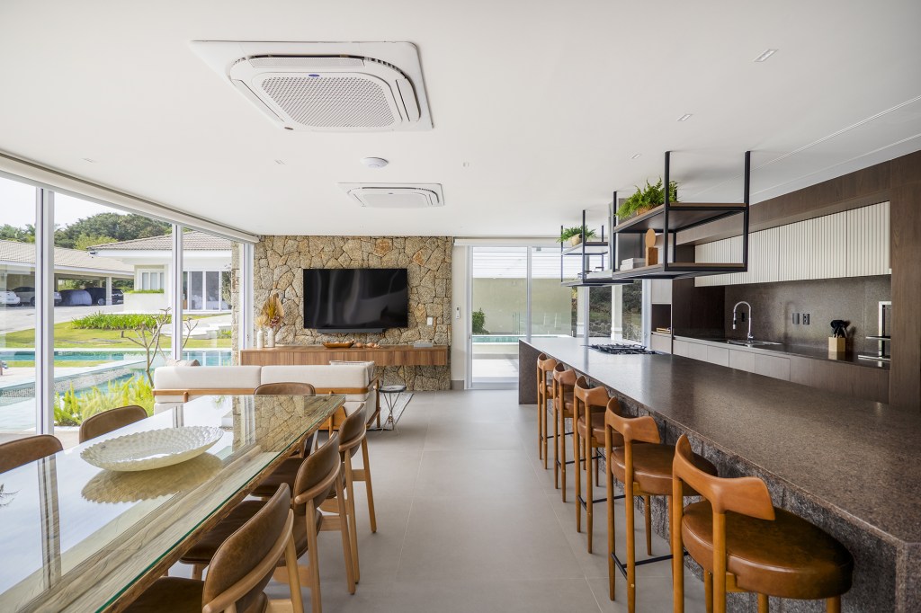 Área gourmet integrada ao jardim é o coração desta casa. Projeto de TM Arquitetura. Na foto, sala de estar na varanda e parede de pedra. Cozinha com ilha.