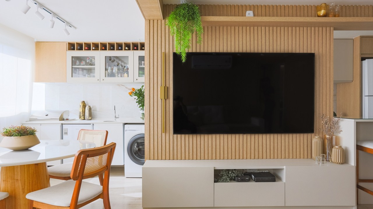 Bege, móveis planejados e muita madeira compõem o décor de apê de 95 m². Projeto de Thais Aguiar. Na foto, sala com varanda e lavanderia. Painel ripado de TV.