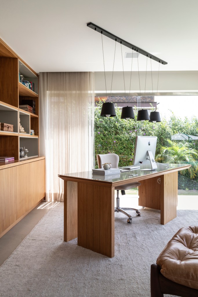 Atemporal e elegante: casa tem lareira e cozinha com piso de ladrilho. Projeto de Carolina Gava. Na foto, home office com estante, mesa de madeira, poltrona Mole.