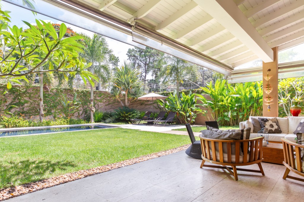 Casa com piscina e varanda gourmet tem sala íntima para videogames e jogos. Projeto de Dantas & Passos Arquitetura. Na foto, varanda externa, poltronas, sofá, jardim ao fundo.