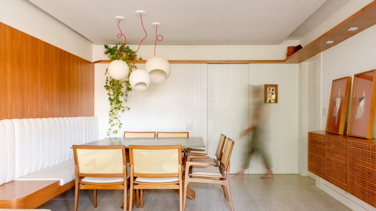 Madeira em tom natural e pintada de branco dão leveza à apê de 117 m². Projeto de Mariana Monnerat e Tainá Binato. Na foto, sala de jantar com mesa quadrada, banco com estofado branco no encosto, luminária, prateleira com planta suspensa.