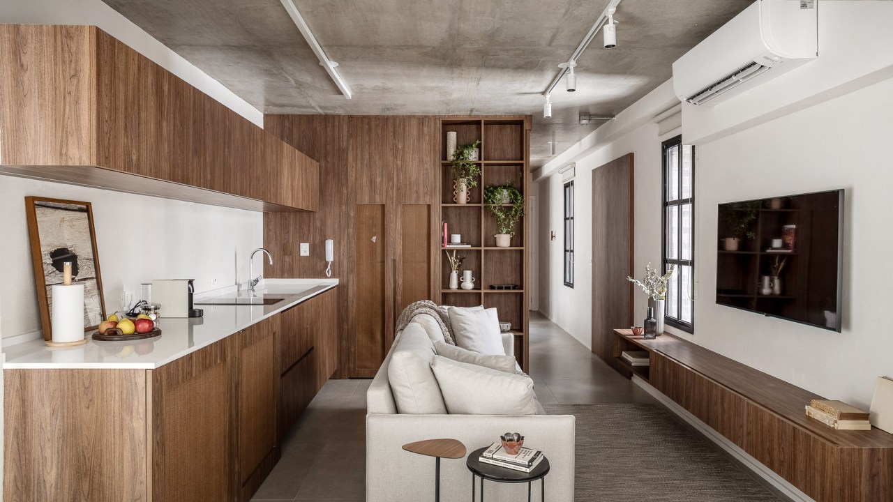 Marcenaria personalizada otimiza espaço e esconde eletros em apê de 48 m². Projeto de Carolina Gava. Na foto, sala integrada com cozinha, sofá branco, piso de porcelanato.