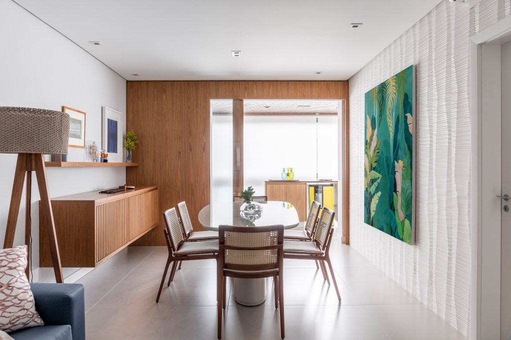 Porta de correr em madeira e vidro separa jantar de varanda neste apê. Projeto de Andrea Bastelaire. Na foto, sala de jantar com piso de porcelanato, mesa de jantar com tampo branco.