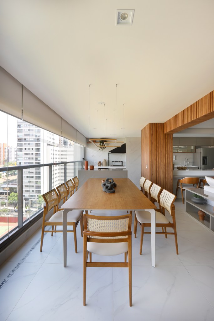 reforma-ape-184-m-elimina-banheiro-ampliar-suite-duno-arquitetura-17-varanda Reforma em apê de 184 m² elimina banheiro para ampliar suíte. Projeto de Duno Arquitetura. Na foto, varanda integrada com mesa grande de madeira, área gourmet ao fundo.