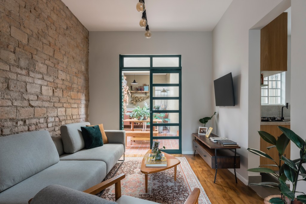 Sobrado de 100 m² ganha solarium e décor que é mix de rústico e retrô. Projeto de Palladino Arquitetura. Na foto, sala de estar com sofá, mesa de centro, tapete, parede de tijolinhos.