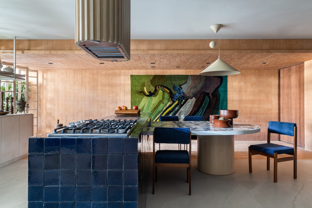 Cerâmicas artesanais compõem o tampo da mesa desta cozinha repleta de azul. Projeto de Mandril Arquitetura. Na foto, cozinha com bancada azul, mesa de azulejos e tijolinhos.