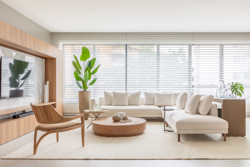 Biblioteca em sala de estar reflete paixão por leitura em apartamento. Projeto de NK Arquitetura. Na foto, sala, tapete bege, sofá branco, planta.