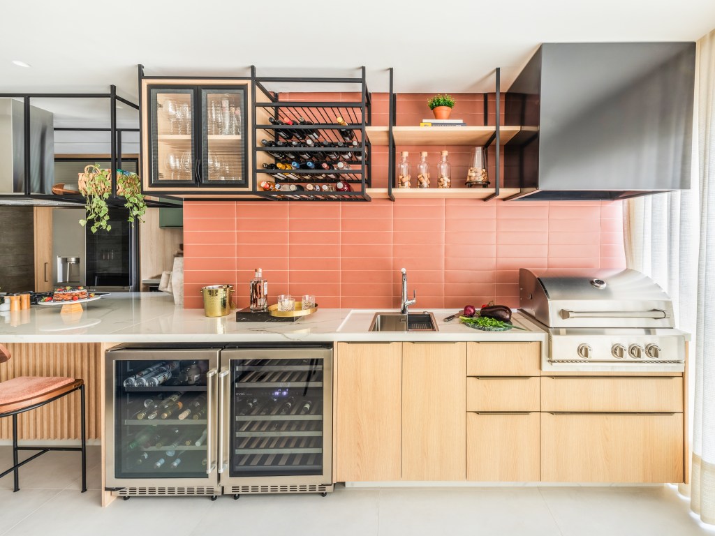 Bancada de lâmina sintetizada integra cozinha e área gourmet neste apartamento. Projeto de BMA Studio. Na foto, cozinha, backsplash de azulejos peach fuzz, salmão, prateleiras aéreas.