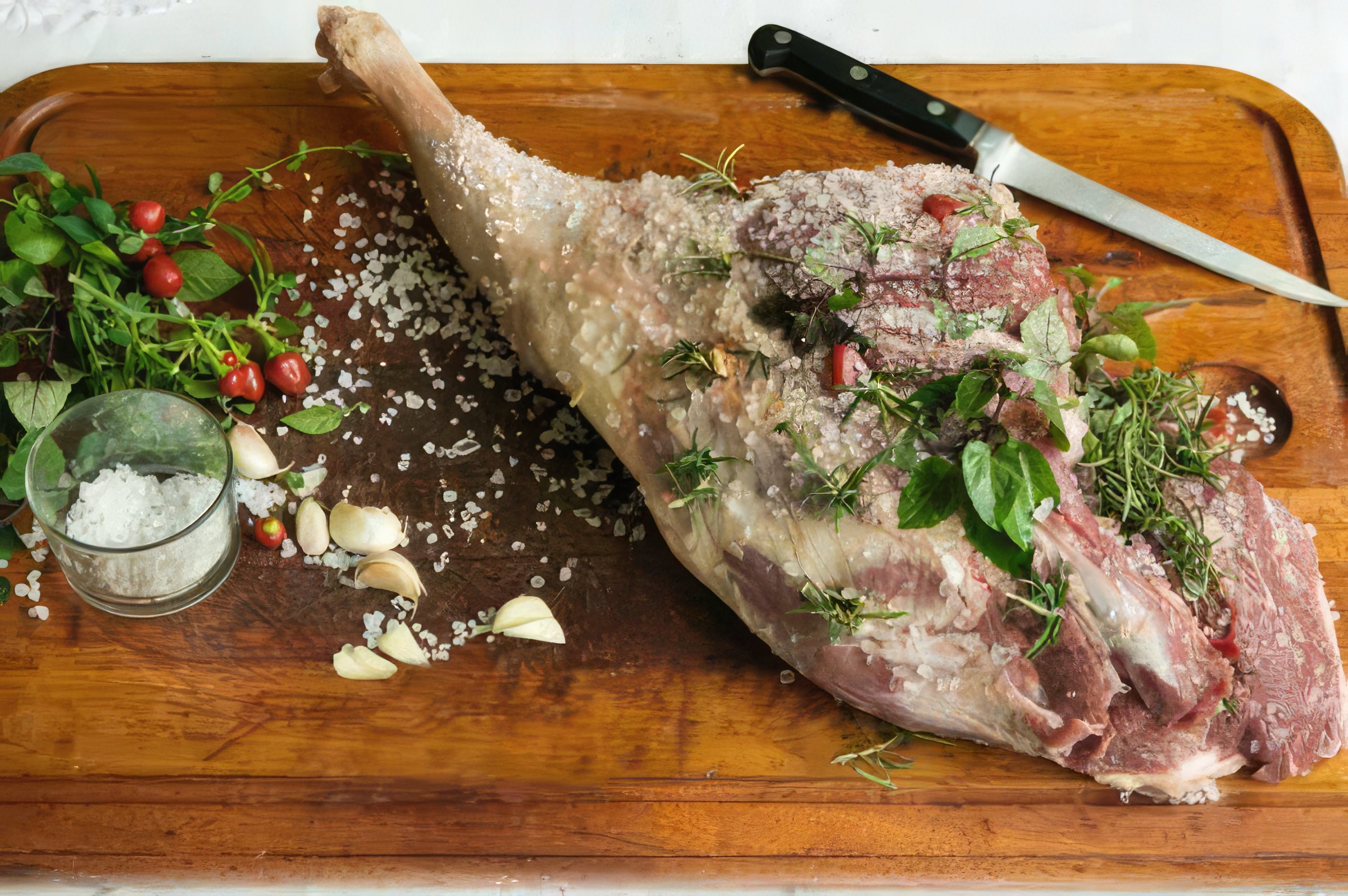 A imagem mostra um delicioso pernil de cordeiro sendo assado na churrasqueira, com uma crosta dourada e suculenta, cercado por chamas e brasas, o que indica que foi bem temperado e est&aacute; no processo de adquirir um sabor defumado.