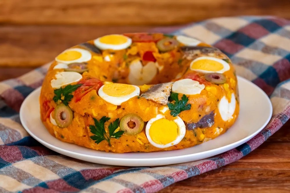 Cuscuz paulista em formato de anel, com ovos cozidos fatiados, azeitonas verdes, sardinha e salsa, sobre prato branco e toalha xadrez